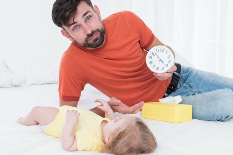 An awake toddler's father is holding a clock showcasing its past naptime for his kid.