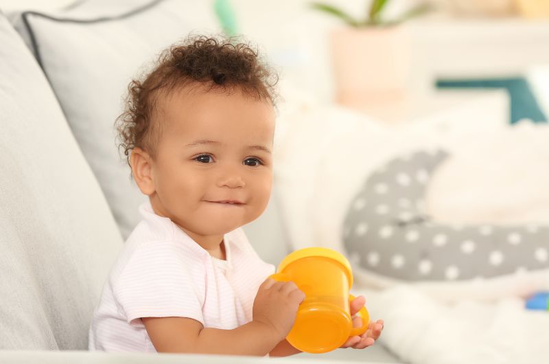 A toddler girl is sitting up and holding her orange sippy cup
