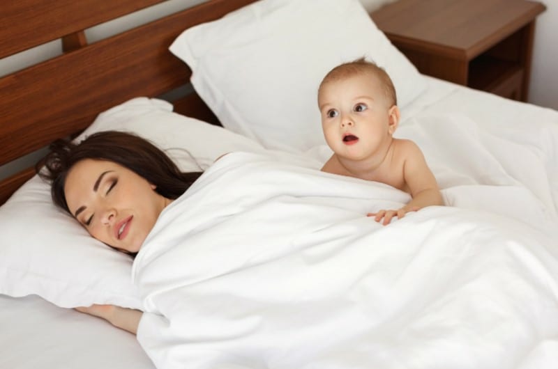 A toddler is awake beside their sleeping mother on a large bed.