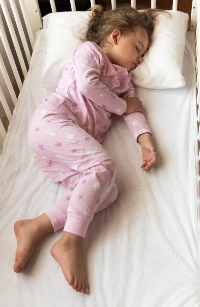 A toddler girl is sleeping in her crib, which has a mattress wrapping cover to protect her from SIDs