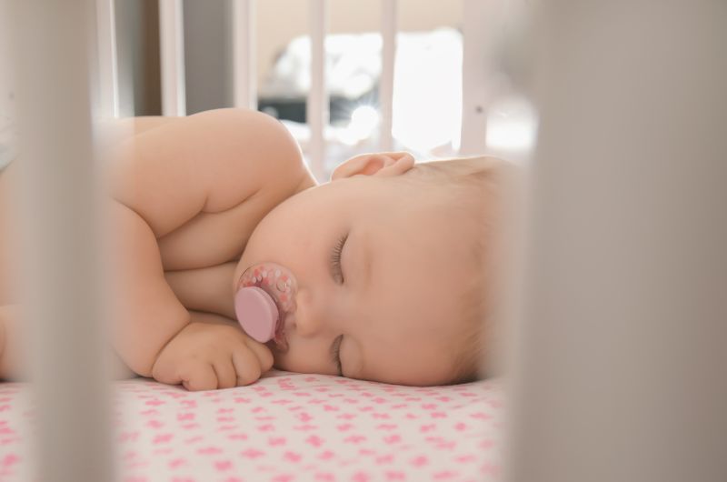 An infant baby is sleeping in her crib with a pacifier in her mouth