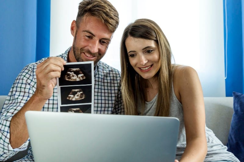 A young couple is showing an ultrasound of their baby to the video chat on the laptop.