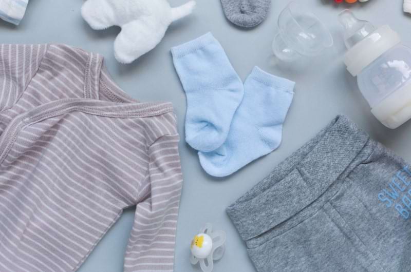Cotton baby clothes including pants, onesie, and socks along with a binkie, and a bottle of milk. 