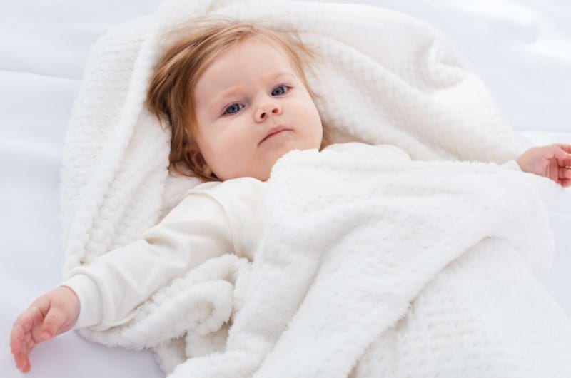 A baby lying on the bed covered in a lightweight blanket.