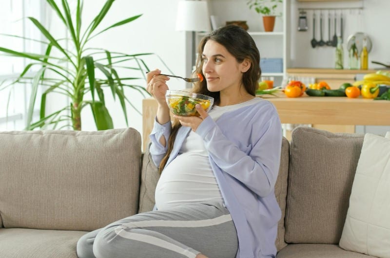 A pregnant woman is sitting on the couch eating a healthy, fiber-rich meal to help with her constipation. 