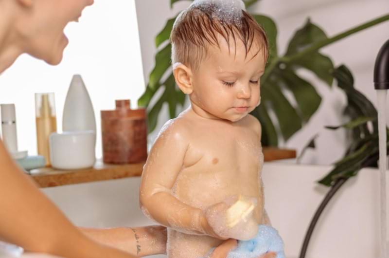 Baby is standing in the tub covered in soapy water as mother gives him a bath.
