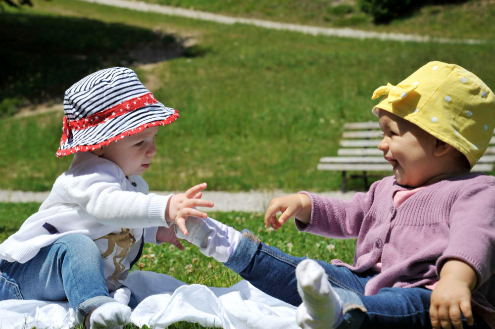 Two toddler girls are sitting on a blanket outside at the park, playing and interacting with each other.