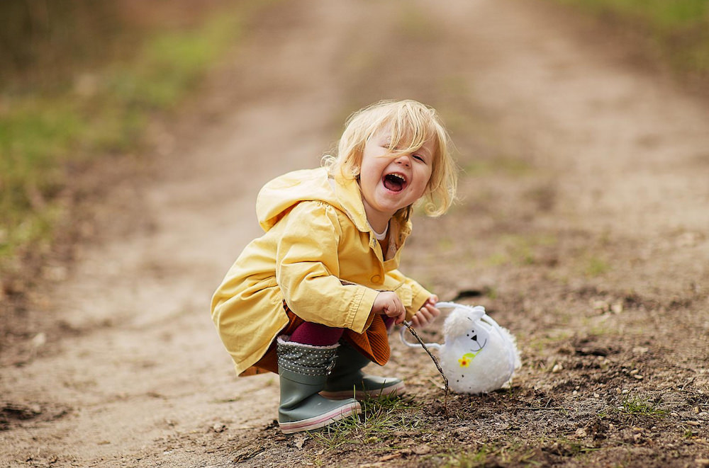 A toddler girl has her boots on and is playing with the dirt and mud outside on a dirt road