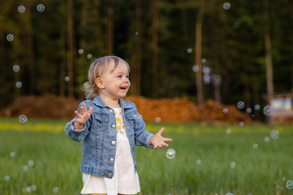 A toddler girl is happily running around outside and popping soap bubbles that are floating in the air
