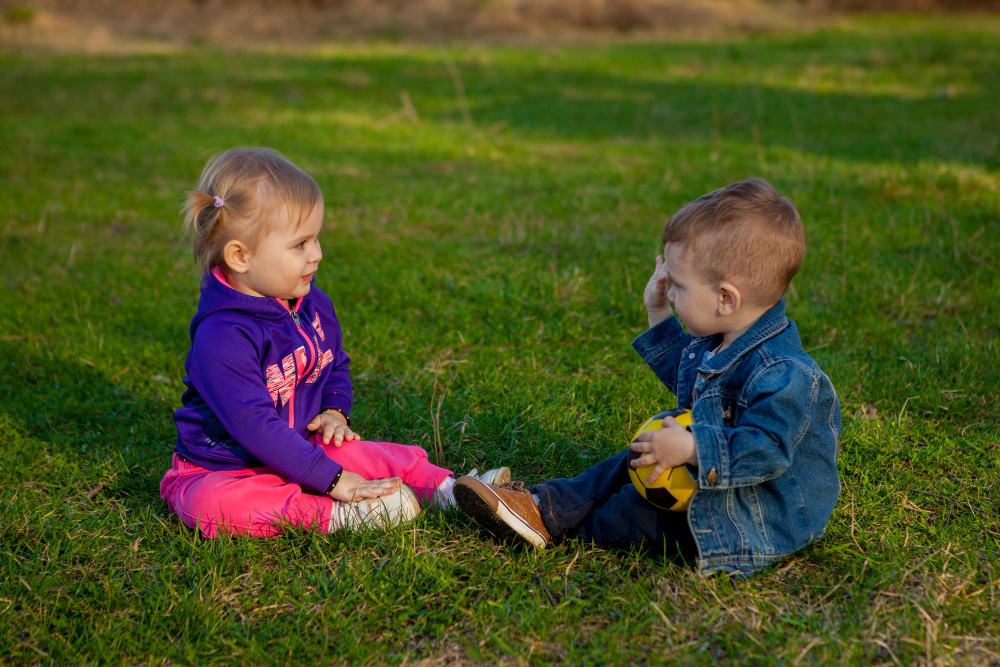 A toddler girl and boy are facing each other and playing outside on the grass