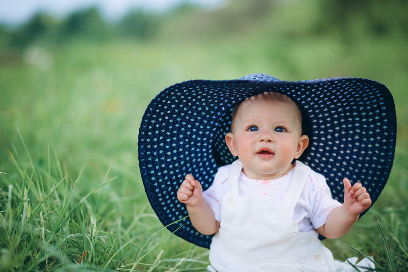 A cute infant girl is sitting on the grass outside and wearing a large womens hat