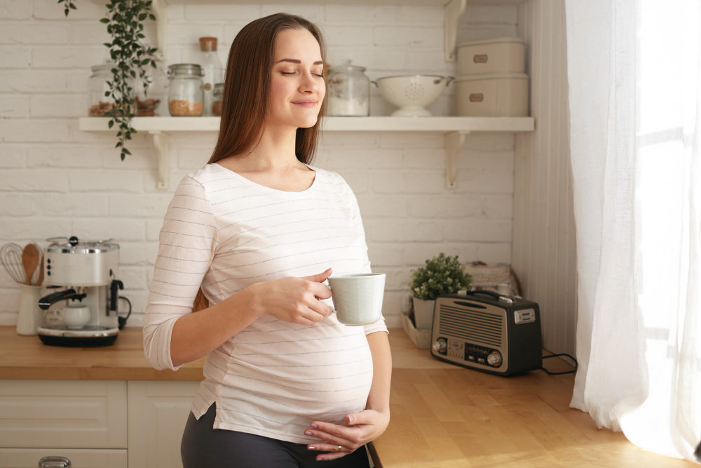 A happy pregnant woman is drinking coffee in her kitchen