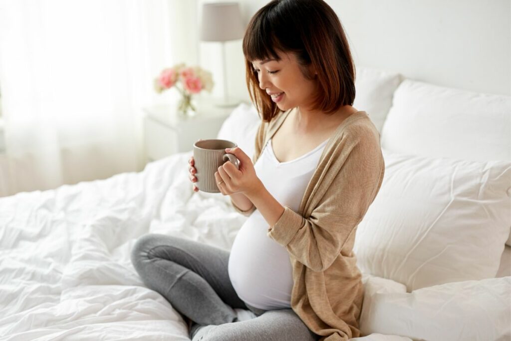 A pregnant woman is happily drinking tea while sitting on her bed on a sunny day