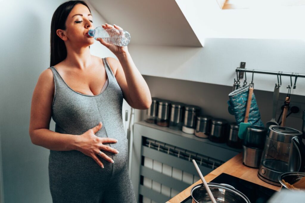 A pregnant woman is drinking water while shes making something in the kitchen