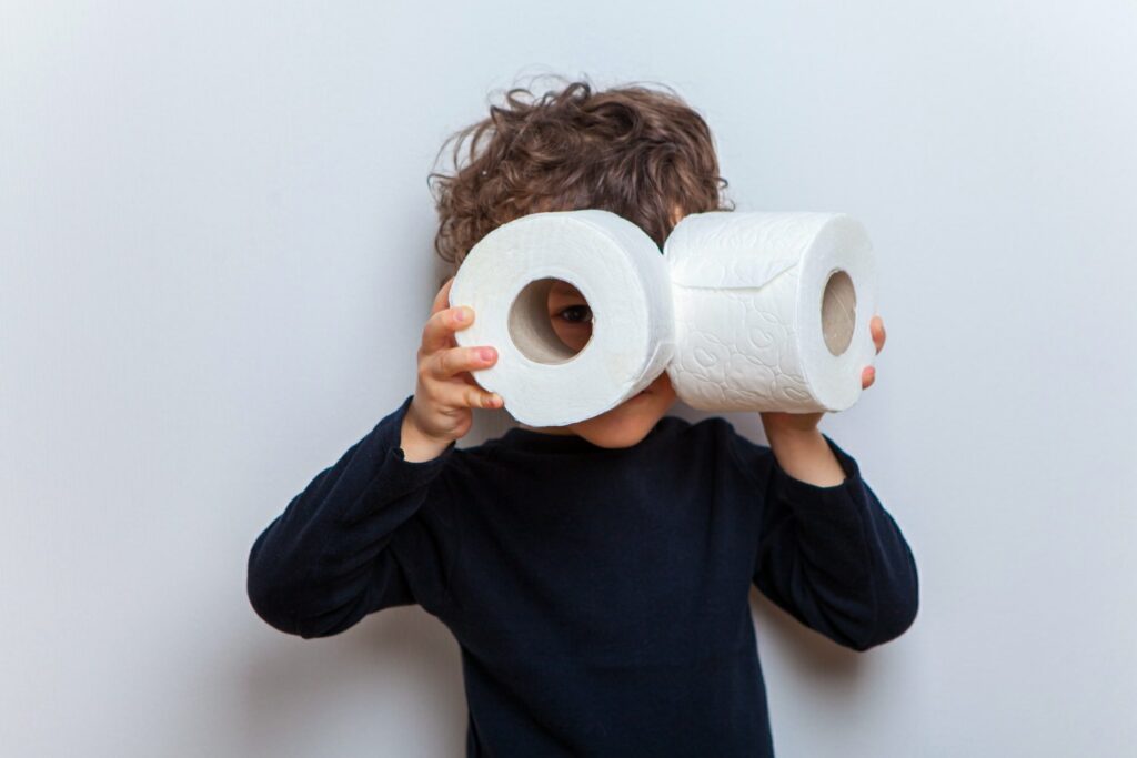 A toddler boy is being silly and is holding 2 toilet paper rolls by his eyes as if they were goggles