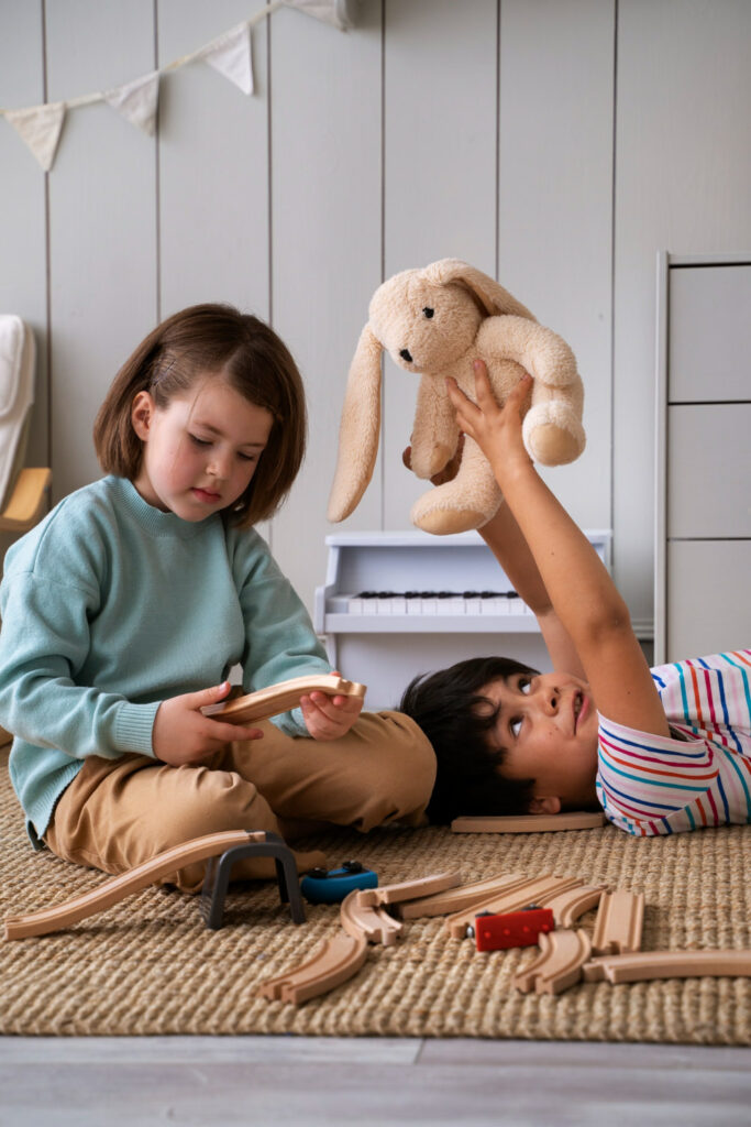 A toddler boy is playing with his stuffed bunny while his sister is playing with train tracks