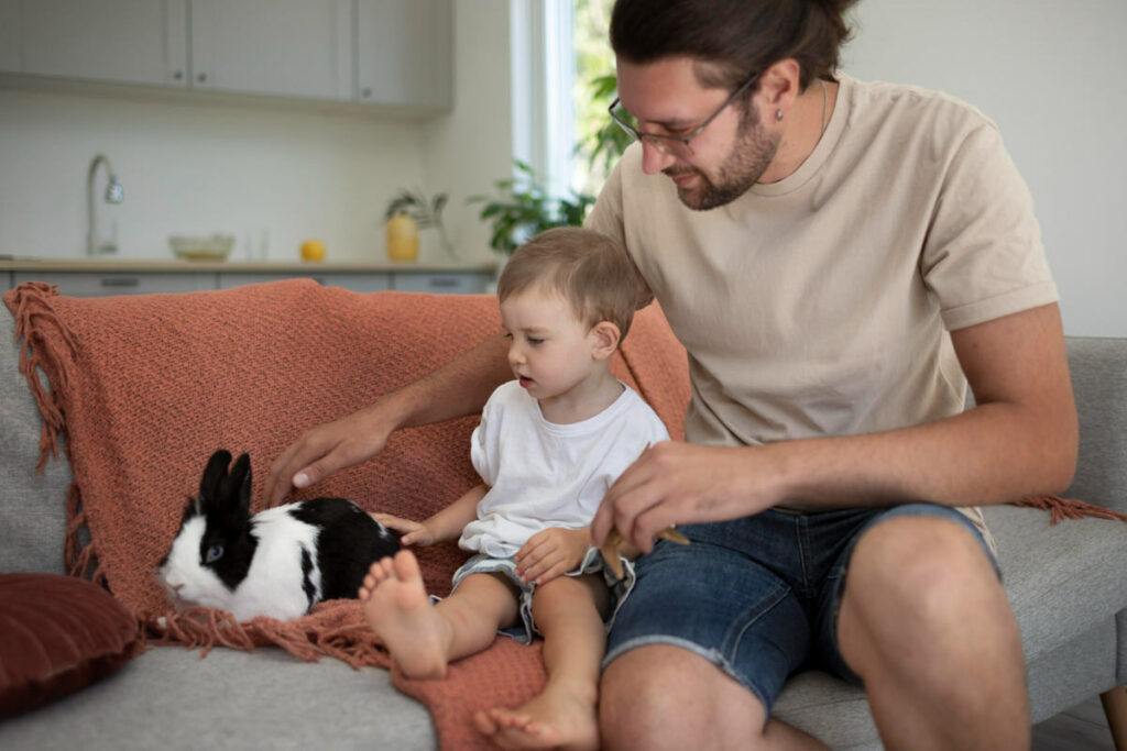 A young dad is sitting next to his toddler son and a cute bunny, and the 2 of them are playing gently with the bunny as a sensory activity for the toddler boy.