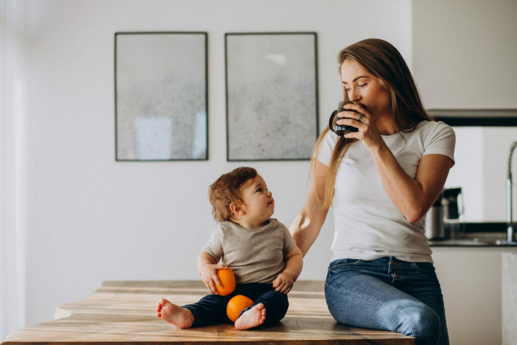 A mom is drinking milk from a cup while sitting on the dining table next to her infant son